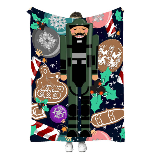 a closeup of the blanket with a cartoon style nutcracker in a bomb suit surrounded by ornaments, holly clusters with old school bombs instead of berries, gingerbread cookies with icing in the shapes of a robot, 1,000lb bomb, and EOD Coin, candy striped dynamite with lit fuzes, and tiny white EOD badges scattered in the background to look like snow on a solid dark navy blue background. Held up by a person to show its size
