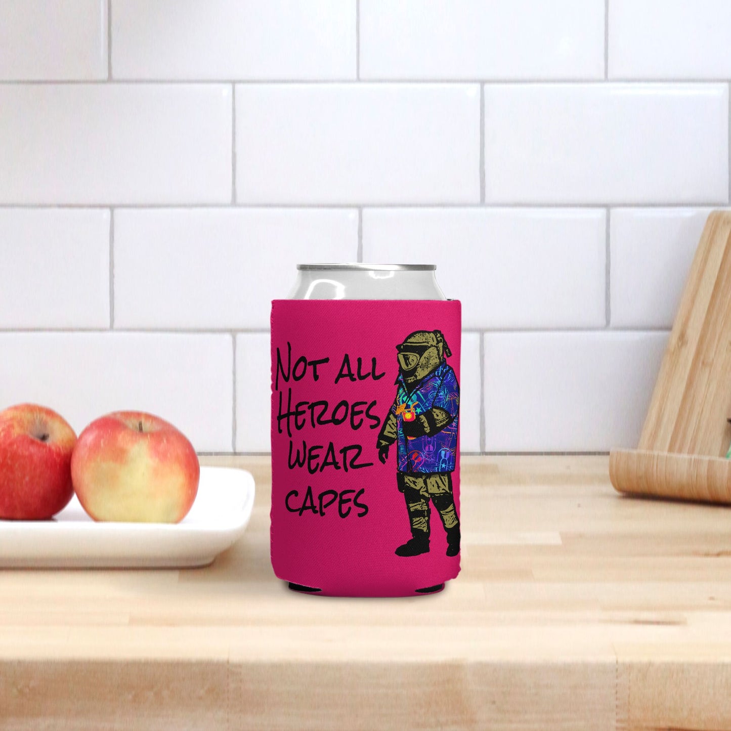 Pink can cooler with superhero design and text on a kitchen counter with apples and a cutting board.