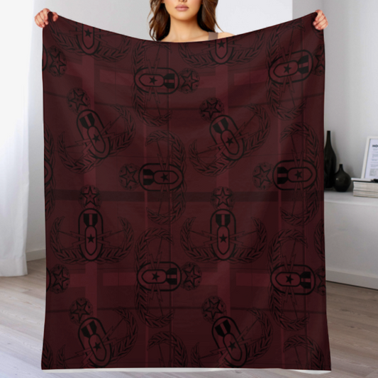 a closeup of throw blanket with black clipart style EOD Master Badges evenly scattered atop ight and dark plaid design on a maroon background Held up by a person to show its size