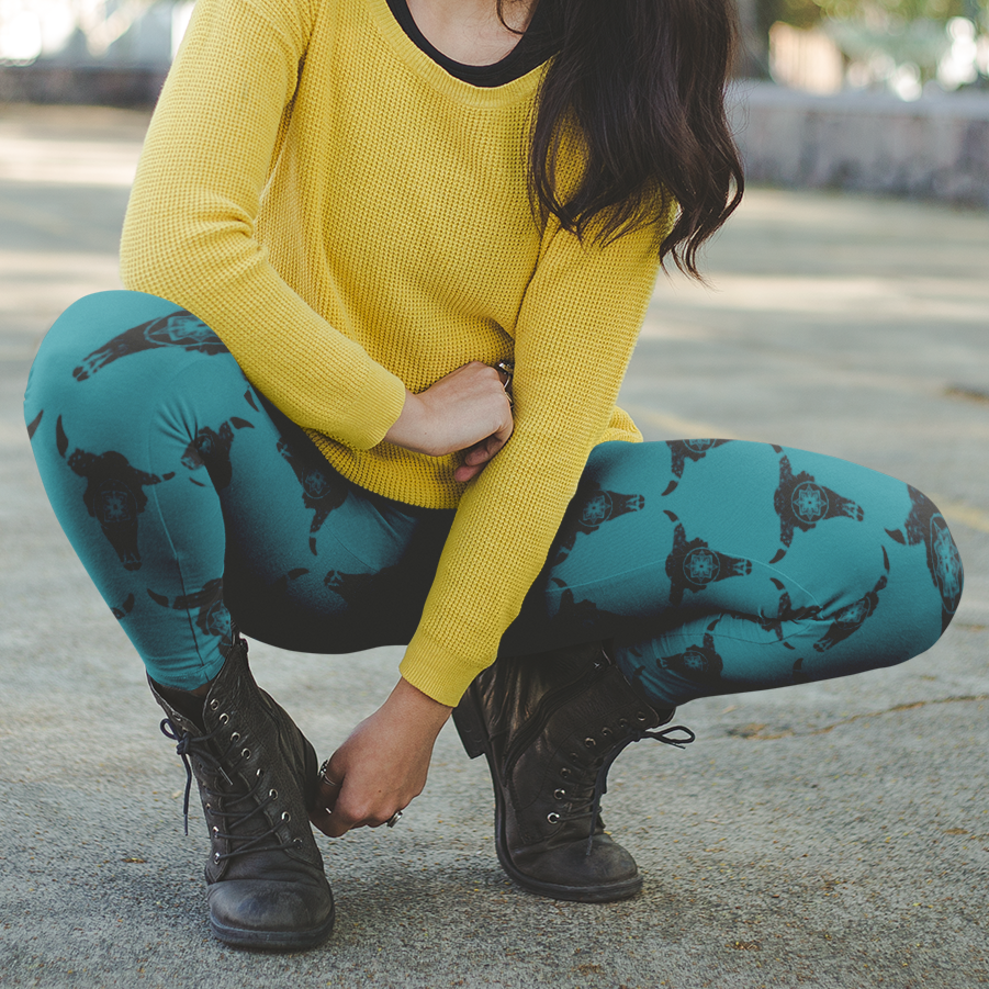 A Woman with dark brown, shoulder length hair, squat down casually in the middle of a road wearing a yellow sweater, teal leggings with black steer skulls pattern and black lace up boots posing for the camera