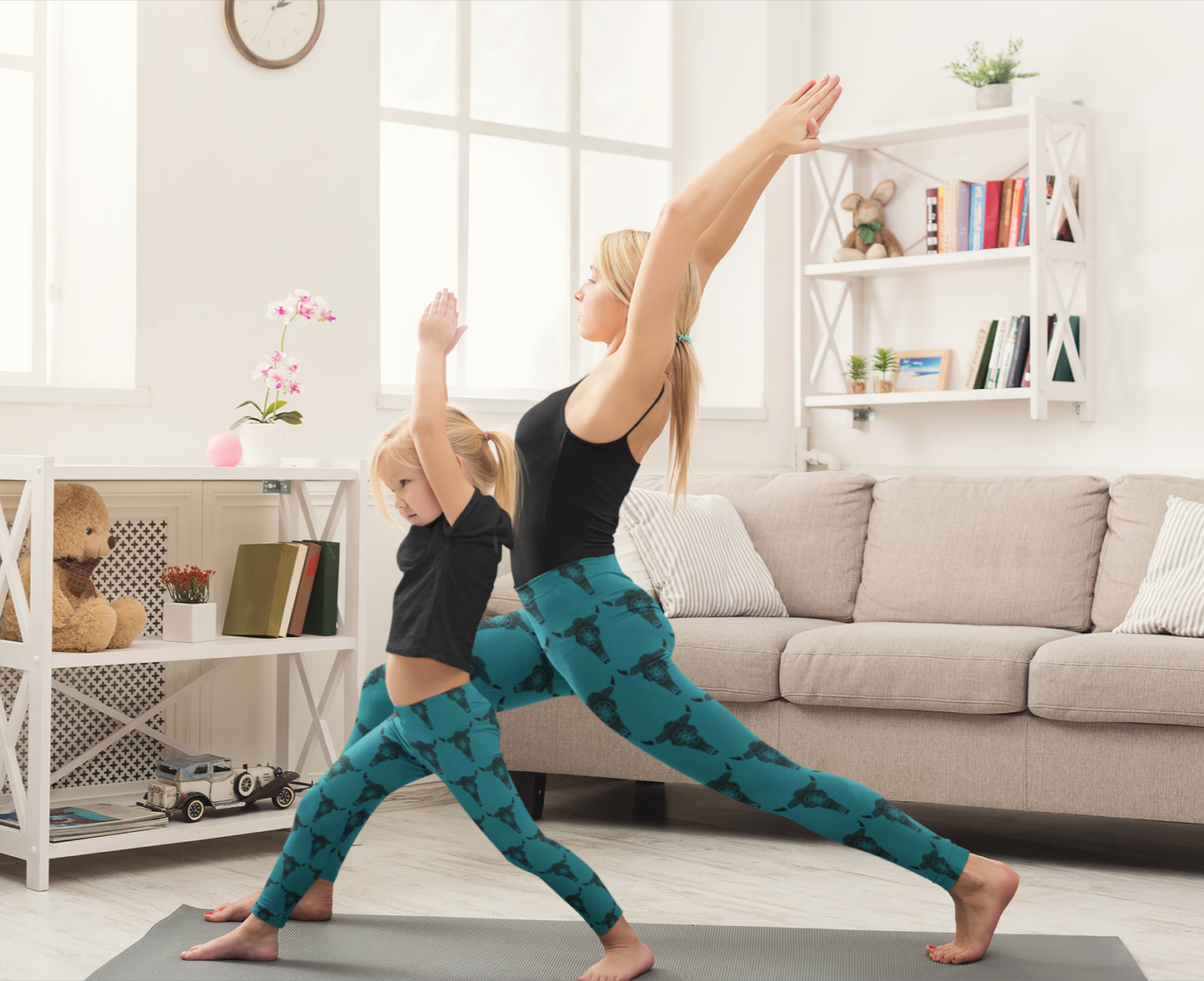 A woman and her daughter doing yoga Warrior pose, wearing high waisted teal yoga style leggings with a black bison skull pattern and black tops in their living room